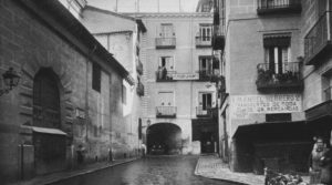 En esta fotografía de la Plazuela de San Ginés en 1931, se puede apreciar al fondo El Arco de San Ginés que da acceso al pasaje del mismo nombre. A la derecha del arco se encuentra la actual Chocolatería de San Ginés, que entonces era una churrería (donde iba de pequeño a comprar para desayunar). A la izquierda la parte posterior de la iglesia de San Ginés.